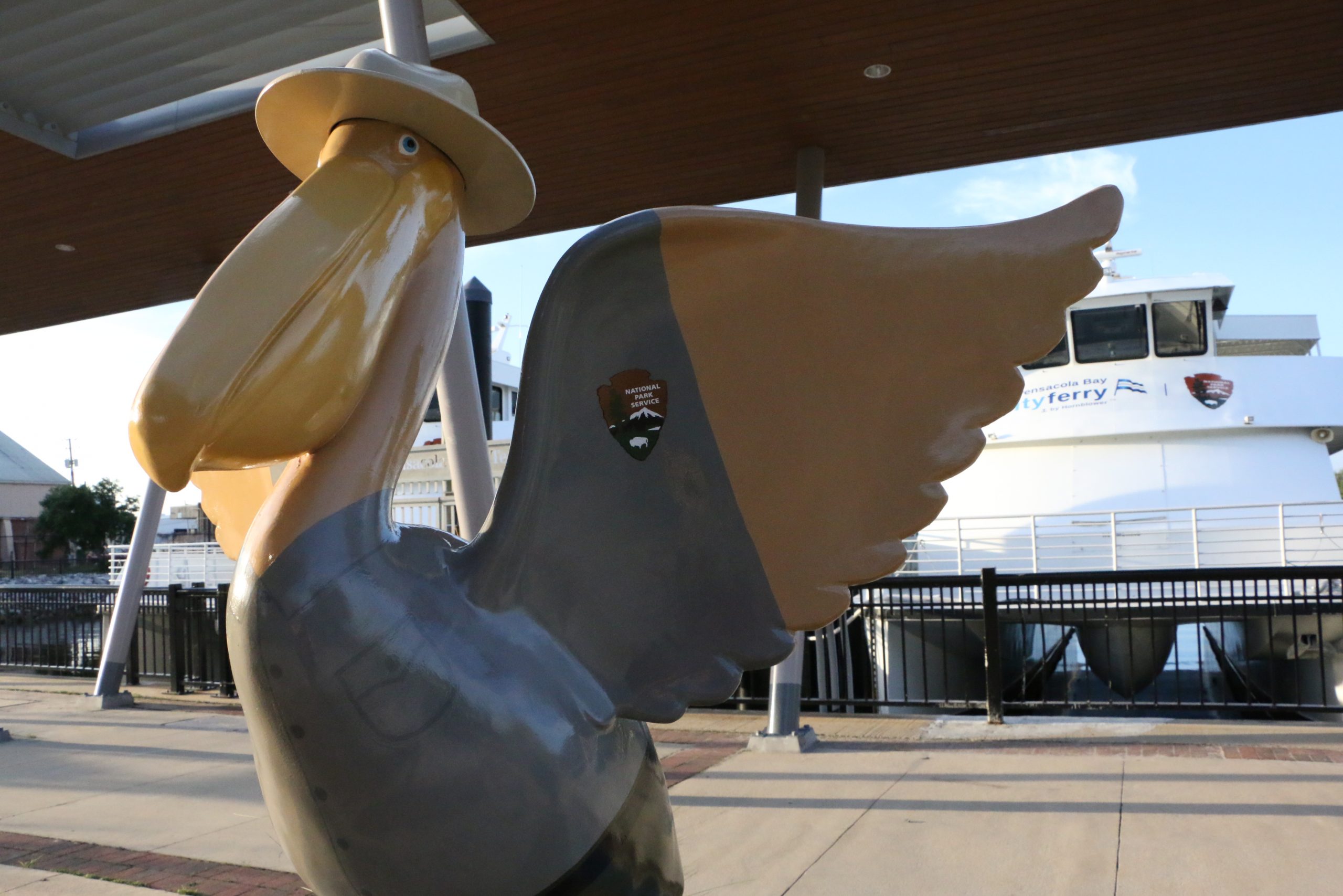 A photo of a pelican statue painted to be national park themed in front of a pier where the Pensacola city ferry is docked.
